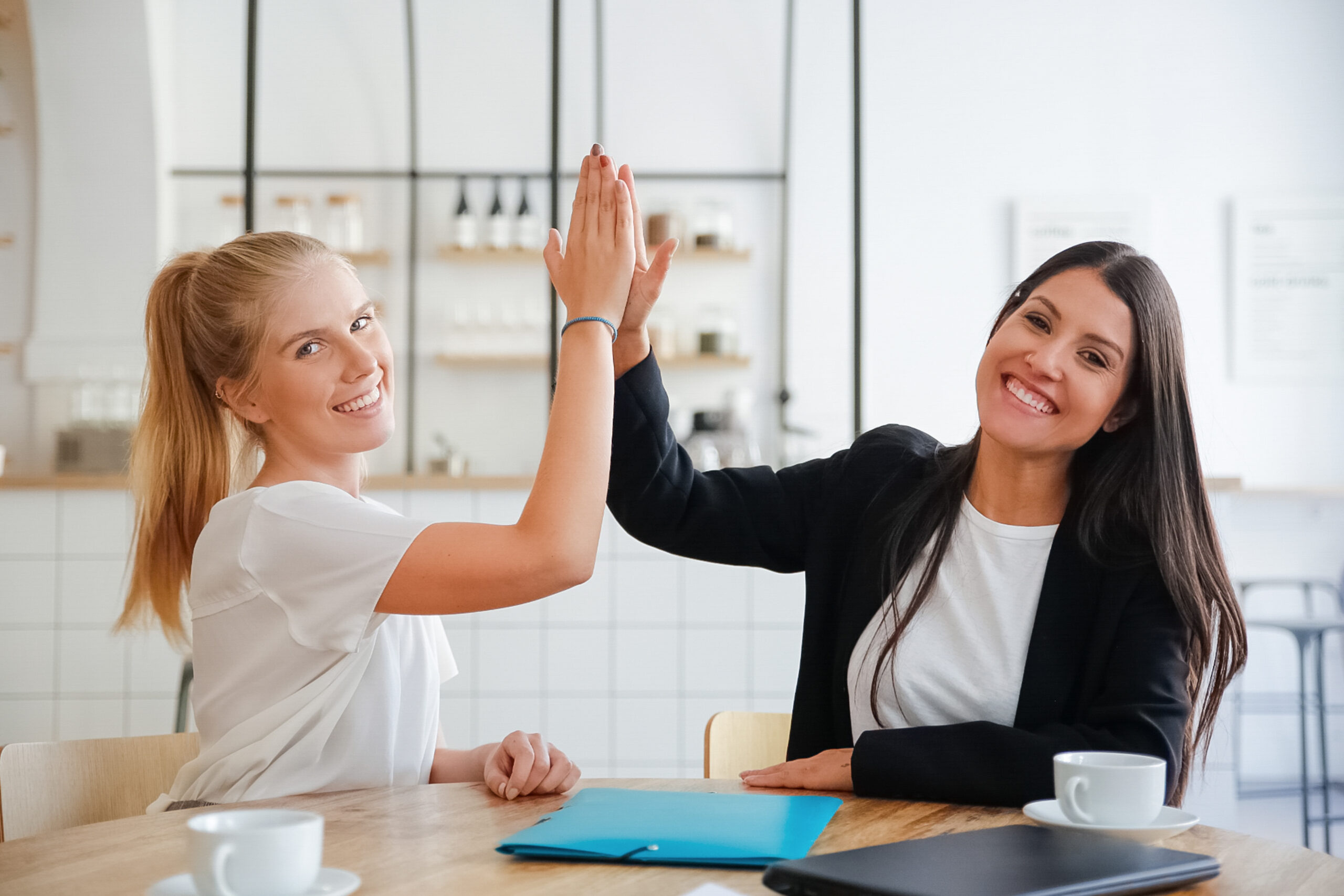 Happy young business women giving high five and celebrating success, sitting at table with documents and coffee cups, looking at camera. Medium shot, front view. Teamwork or cooperation concept
